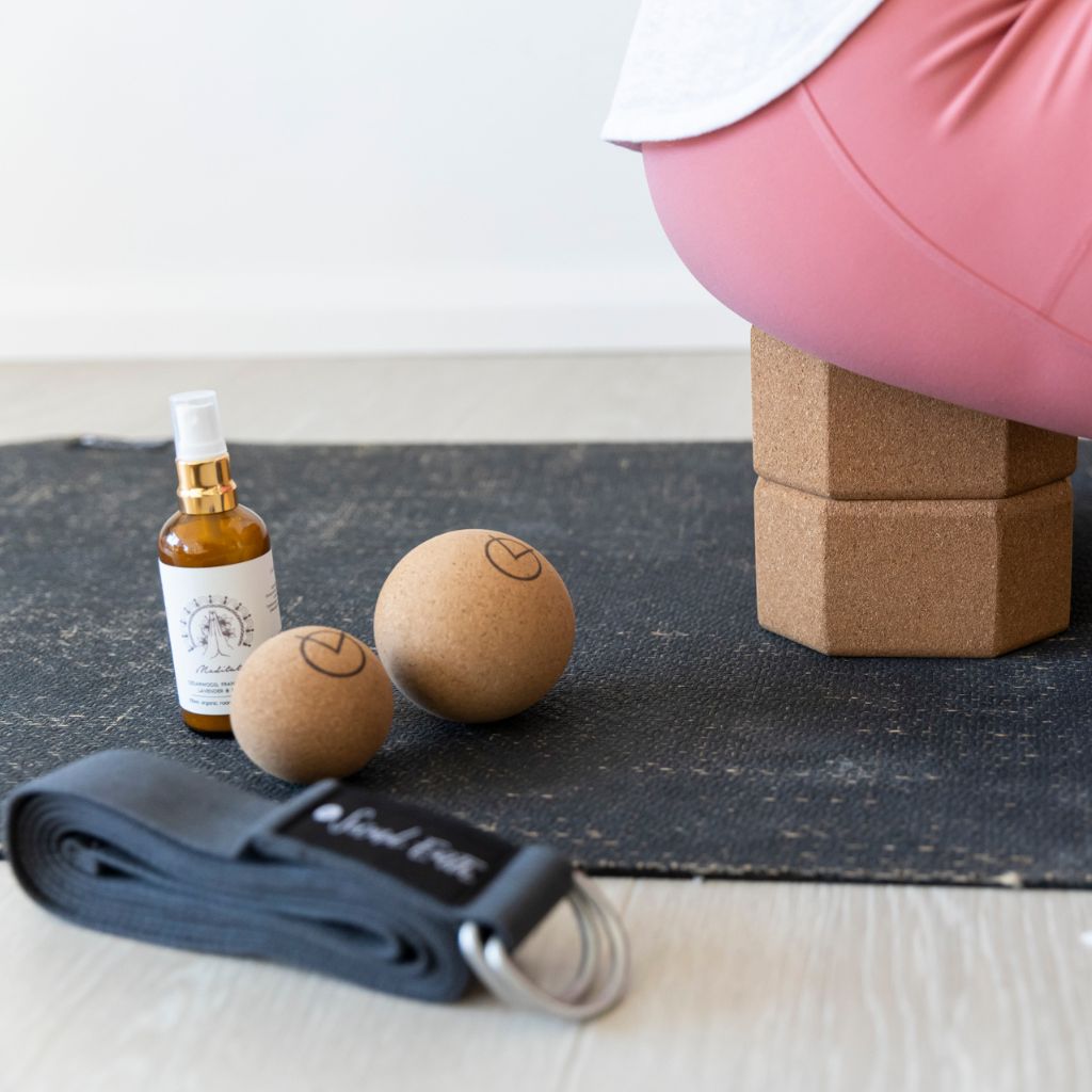 Woman sat on cork yoga blocks laid on black jute eco-yoga mat, with meditation spray, cork balls and a yoga strap next to her, taking time out in the office