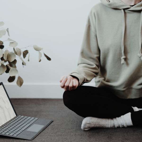 Man in tracksuit meditating cross legged on grey carpet with laptop next to him in his office wellbeing space