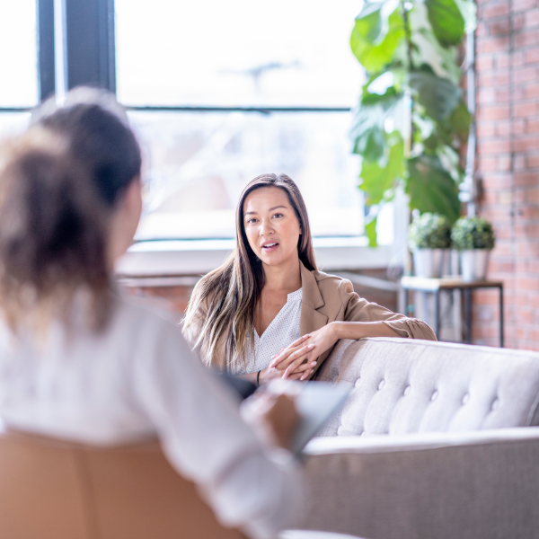 Two women are sitting on a couch and chair in a modern office setting with plants and large windows, having a workplace set-up consultation session