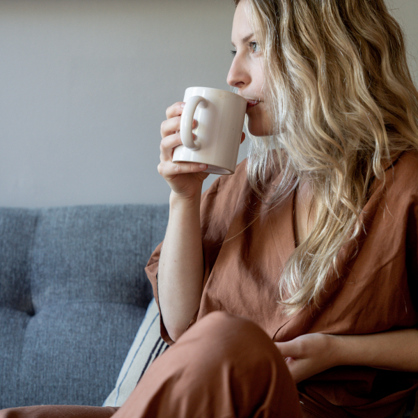 Woman sitting on a couch holding a white mug, wearing a brown outfit, taking some time for herself -  - one of the  tips from her personalised wellbeing consultation sesson