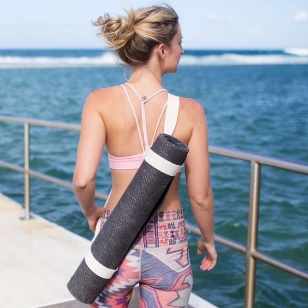 Woman posing on beach pier with rolled black yoga mat slung over shoulders