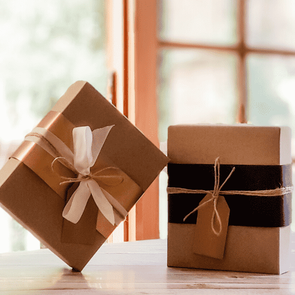 Two Kraft gift boxes leaning on each other on wooden table with gold and black belli bands and Kraft gift tags with blured background of wooden window and trees