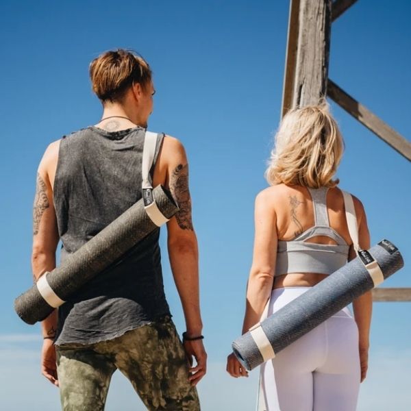 Man and woman standing by wooden beach pier carrying second earth yoga mats over their shoulders