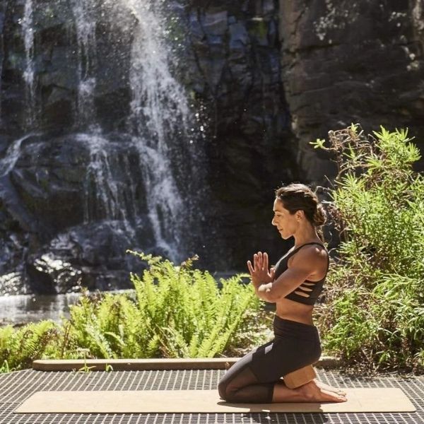 Woman in front of a cliff face with a waterfall sitting with legs tucked under here with a cork Zone yoga rectangular block with corners cut at an angle tcuked between her bottom and feet to give her a straight spine alignment