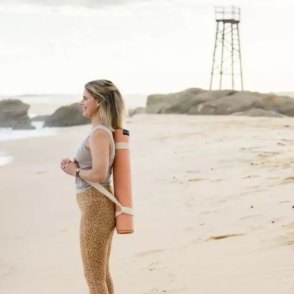 Woman posing on edge of beach with rolled pink clay yoga mat slung over shoulders